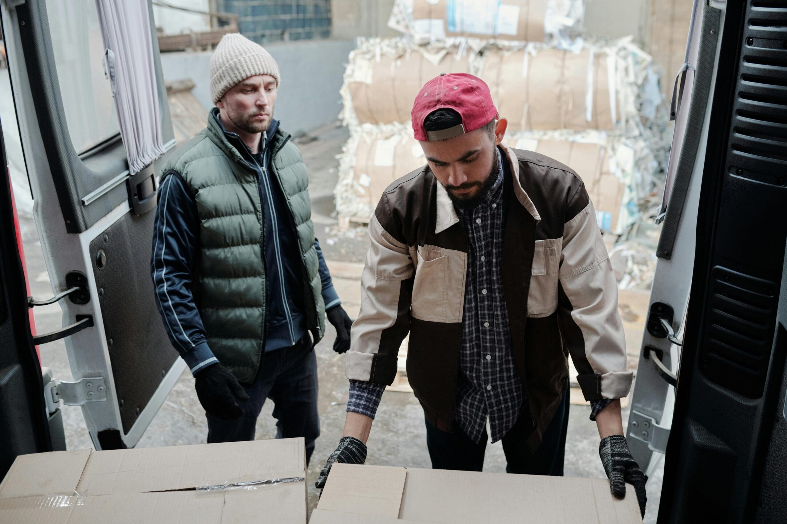 Two men loading cardboard boxes into a delivery van at an urban site.